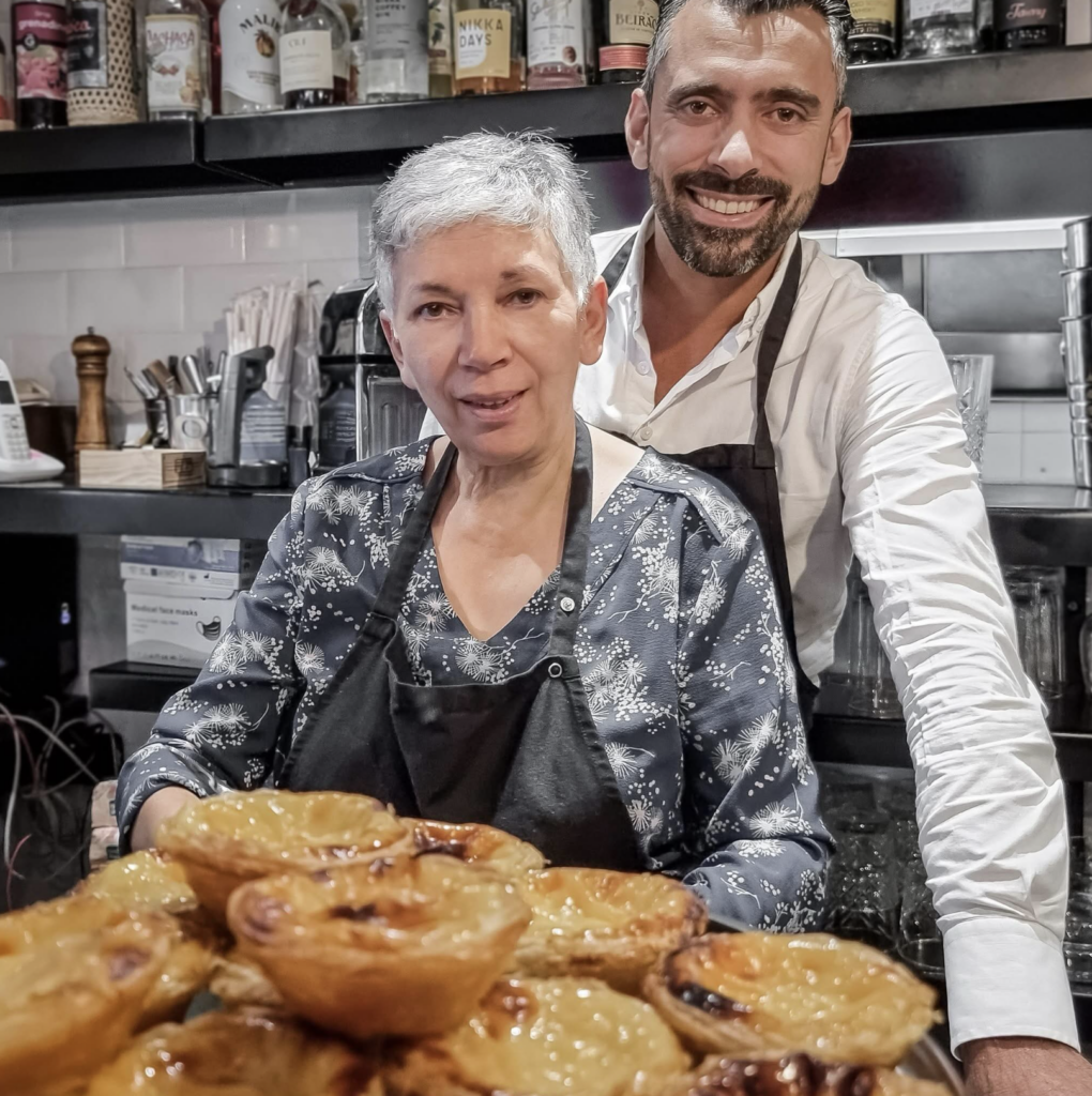 Deux restaurateurs souriants devant des pâtisseries portugaises fraîches dans leur cuisine professionnelle équipée