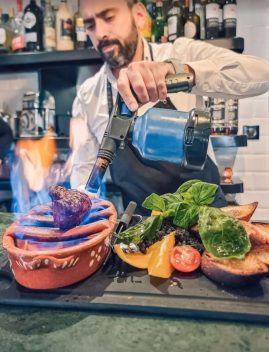 Chef versant une boisson bleue sur des brochettes de viande grillée avec légumes frais dans un restaurant portugais