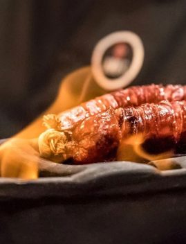 Saucisse grillée portugaise dorée et croustillante dans un plat noir avec flammes, accompagnée d'une rondelle d'oignon